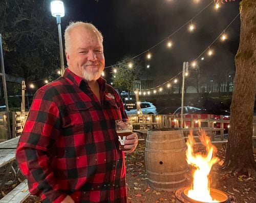 A man in a black and red checkered jacket and holding a glass of beer looks at the camera while outside in a beer garden in front of a fire. Lights are strung overhead, and a wood barrel is in the background.