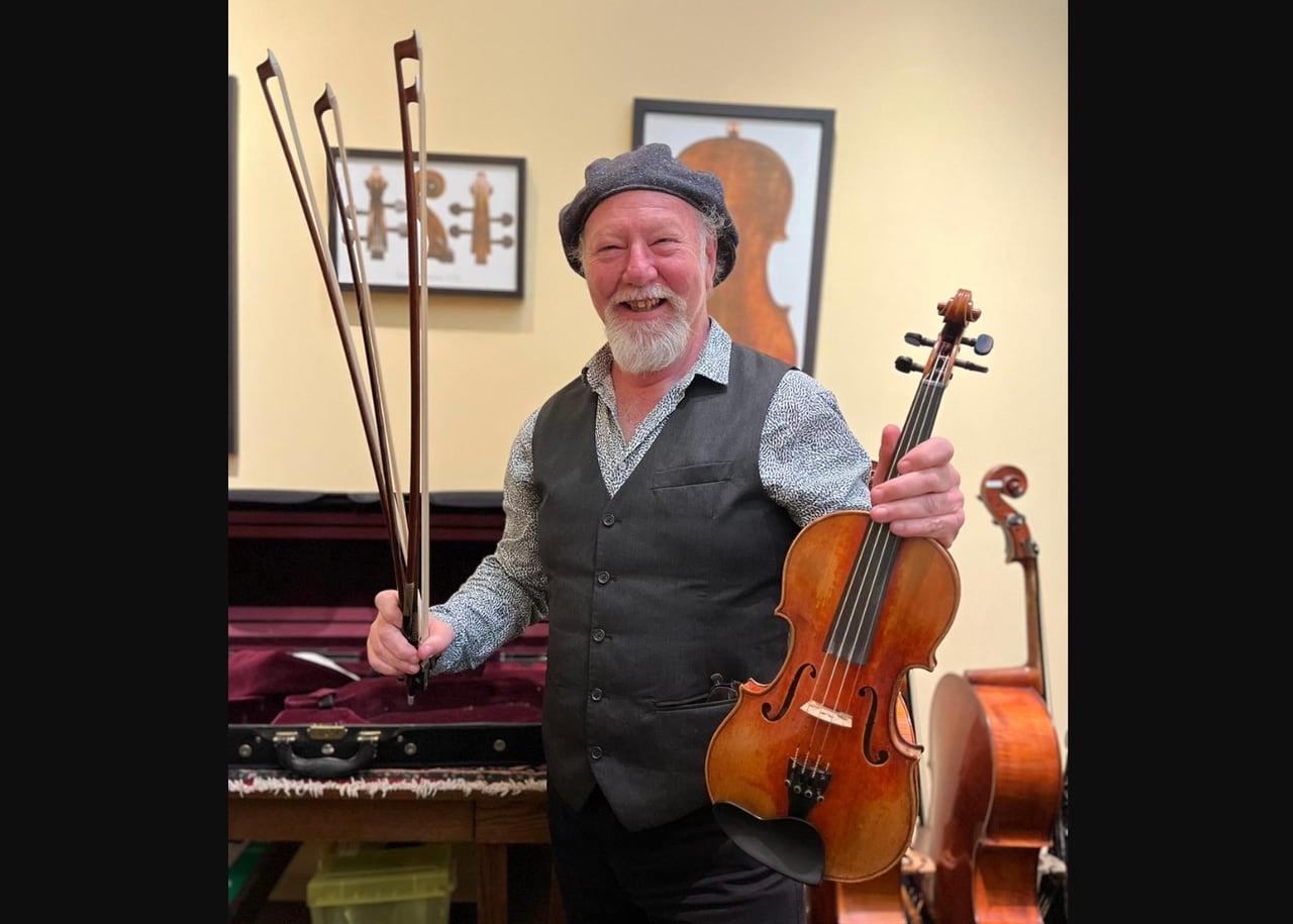A man with a gray beard and a cap holds up a violin and four bows as he smiles widely.