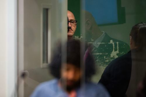 A bald man in glasses peers around a closing door behind a courtroom wall