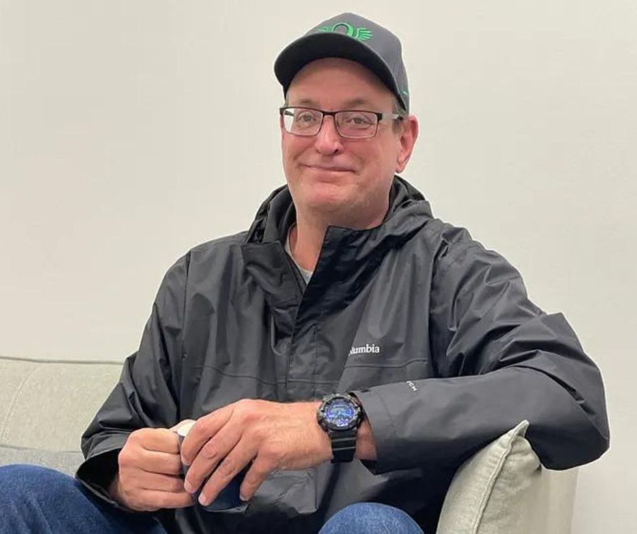 A man in his 50s holds a mug and smiles at the camera. He's sitting down on a white couch against a white wall, wearing glasses, a black baseball cap with a green logo, black jacket and jeans.